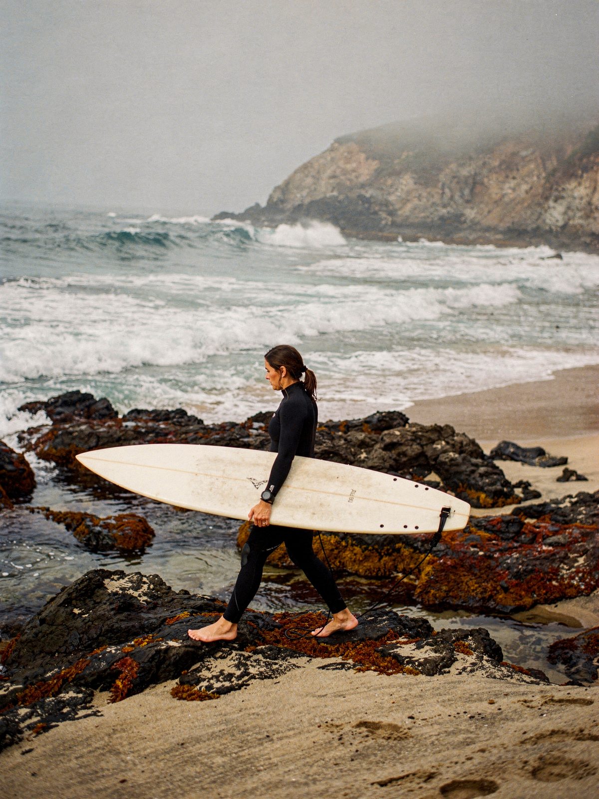 Surfer in a wetsuit carrying a board over rocks toward a foggy point break