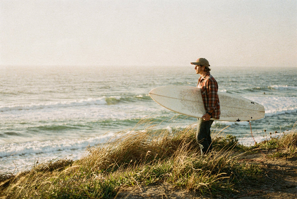 Surfer with a surfboard on a grassy bluff above the ocean at golden hour